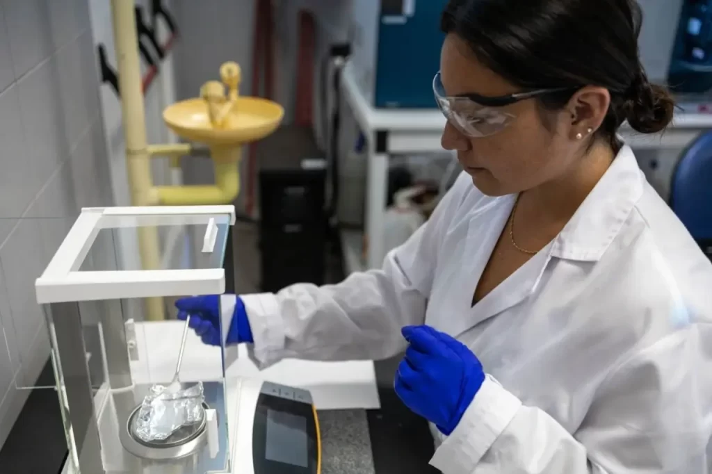 A female scientist in safety goggles a lab coat and blue gloves is using a precision digital scale