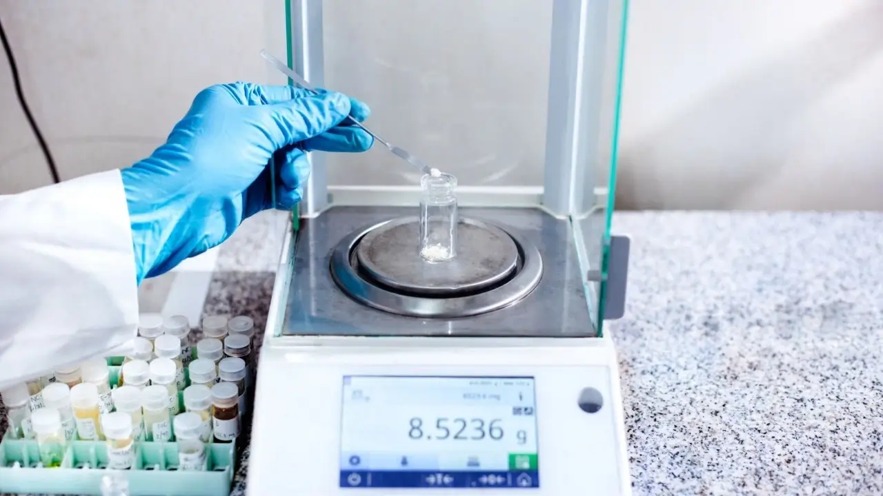 A blue-gloved hand uses a spatula to transfer white powder into a small glass vial on a digital scale, which reads 8.5236g.