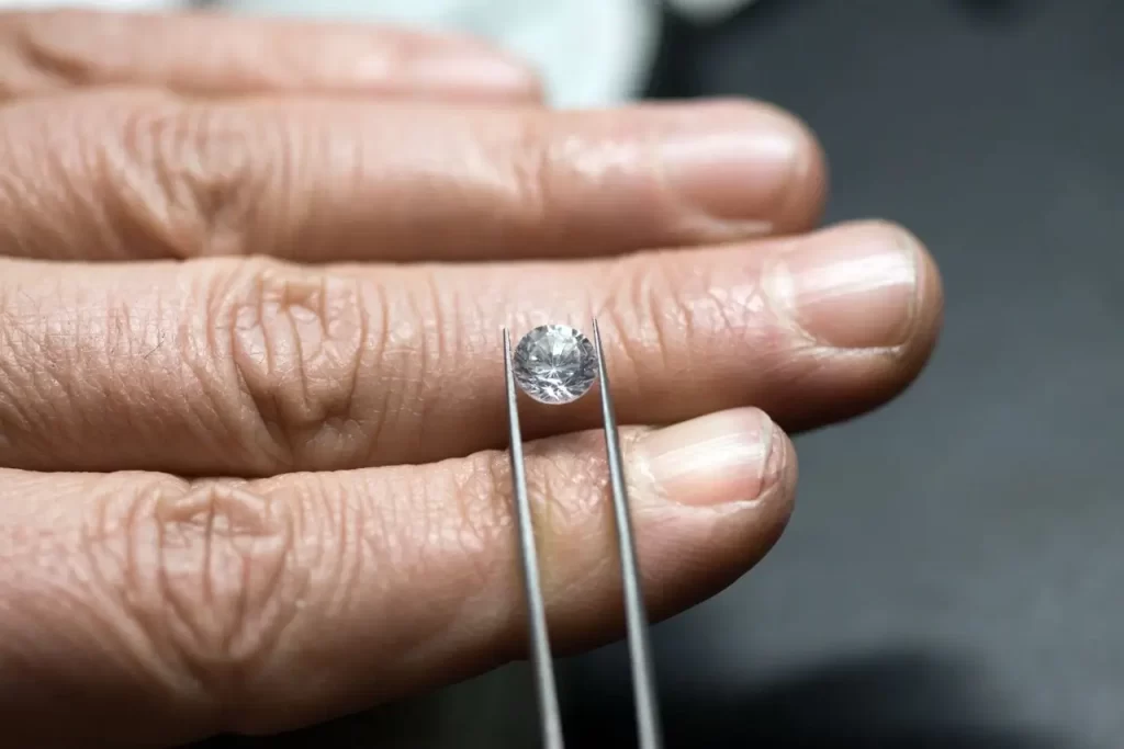 A sparkling diamond is held by tweezers in a close-up view of a wrinkled hand.