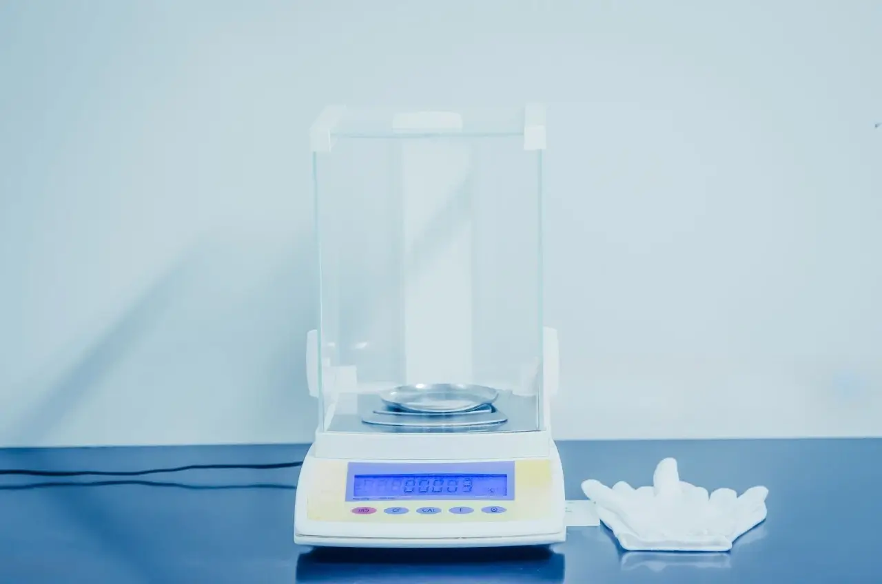 White and yellow high-precision balance on a blue lab bench with a clear shield and glove nearby.