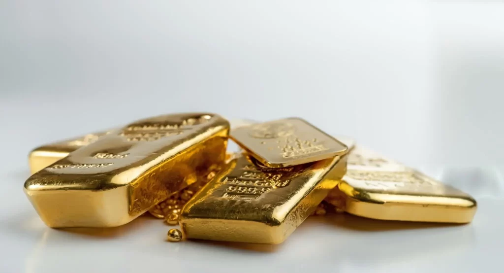 A pile of shiny gold bars and gold pellets rests on a white surface with a bright white background