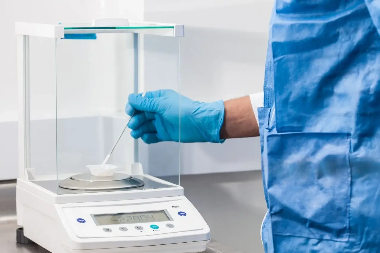 Scientist in blue scrubs using a spatula to add substance to a weigh boat on a high-precision lab balance.