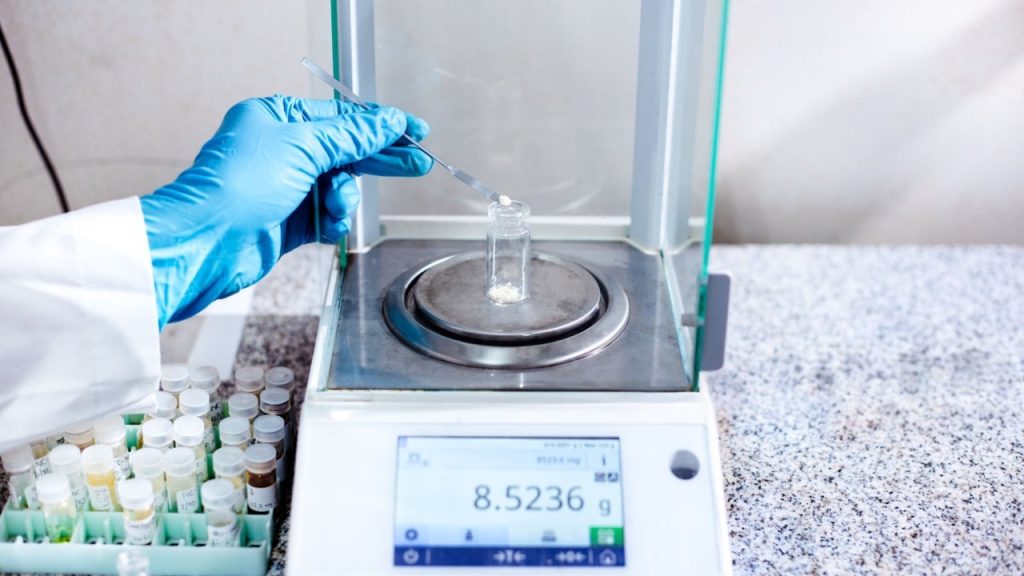 A gloved hand uses a spatula to add white powder into a small glass vial on a digital analytical balance displaying 8.5236 g