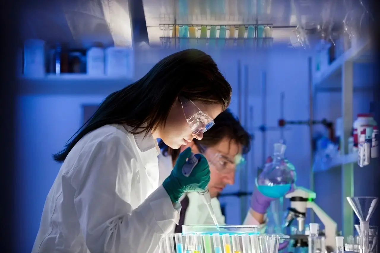 Female scientist in a lab coat safety glasses and gloves using a pipette in a darkly lit busy laboratory setting