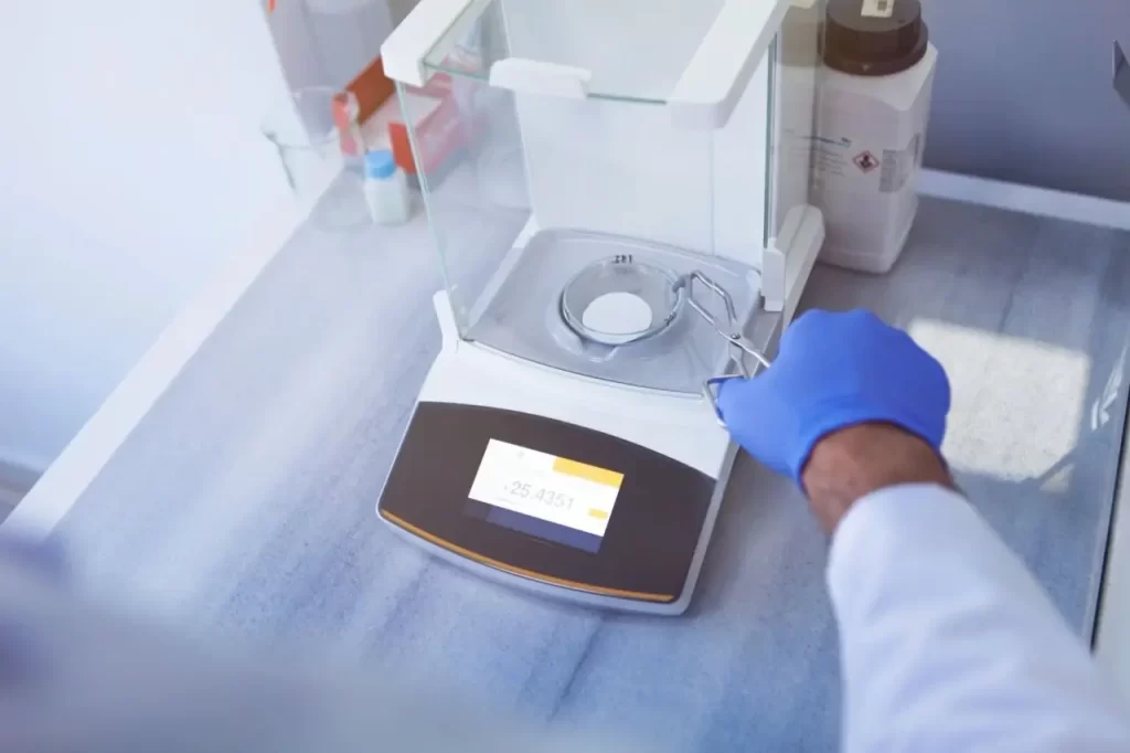Gloved hand with tongs placing filter paper on a lab analytical balance showing a reading of 25.4351.