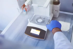 Gloved hand with tongs placing filter paper on a lab analytical balance showing a reading of 25.4351.