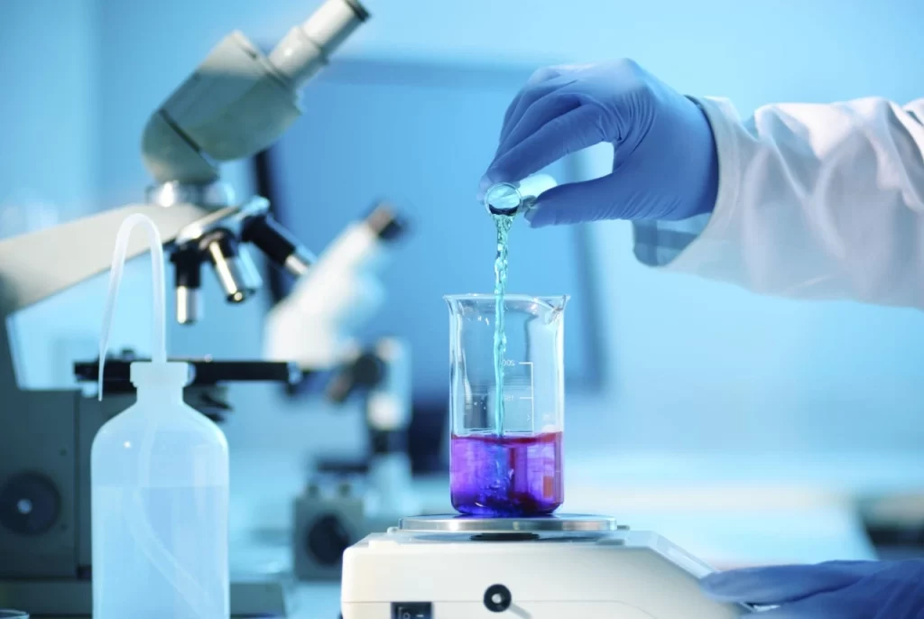 A scientist in blue gloves carefully pouring liquid into a beaker on a digital analytical balance in a lab.