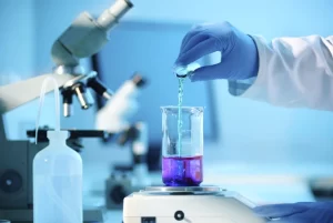 A scientist in blue gloves carefully pouring liquid into a beaker on a digital analytical balance in a lab.