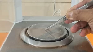 Close-up of a person using tweezers to place a tiny sample on a high-precision analytical balance pan.