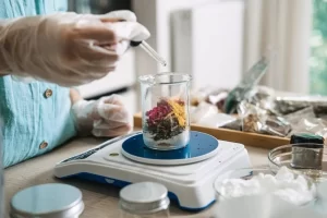 Close-up: A gloved hand uses a pipette to drop liquid into a beaker filled with dried flowers on a white digital scale.