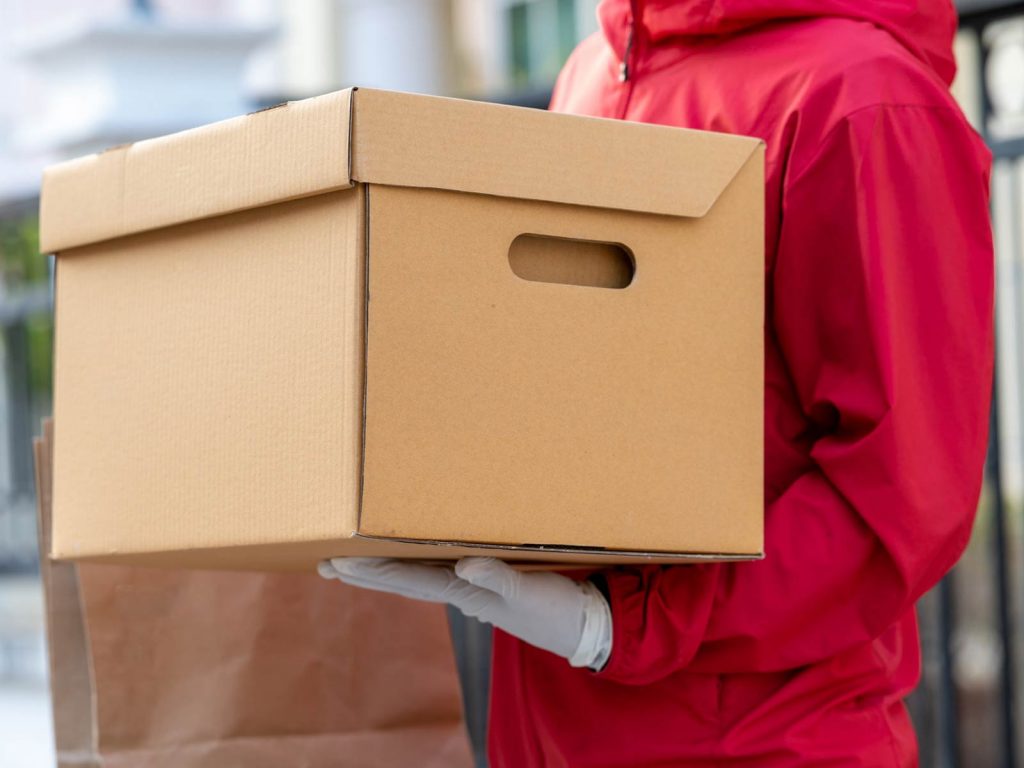 A delivery person in a red jacket holds a large cardboard box with handles, ready to ship a Stuccler lab balance.