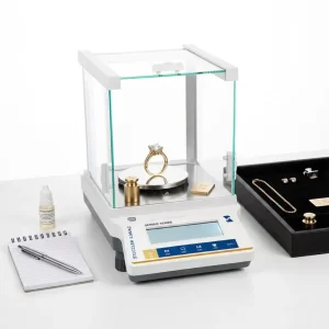 A professional gold balance weighing a diamond ring on a desk with calibration weights and a notebook.
