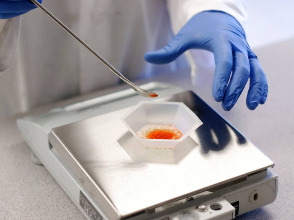 Scientist in blue gloves using a spatula to weigh orange powder on a Stuccler top loading laboratory balance.