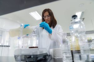 A scientist wearing a white lab coat and safety goggles pouring liquid into a beaker on an electronic balance scale.