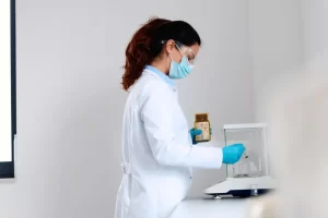 A scientist in a lab coat and mask weighing a sample using a high-precision analytical balance.