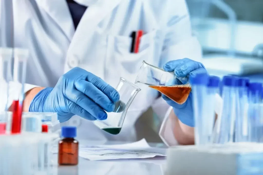 Scientist in blue gloves pouring orange liquid between beakers in a chemical and pharma lab.