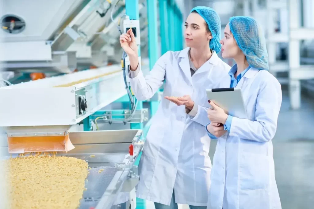 Two QA inspectors in white coats and hairnets check food quality on a manufacturing conveyor belt.