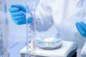 A laboratory technician wearing blue gloves uses a spatula to measure white powder on an electronic balance scale.