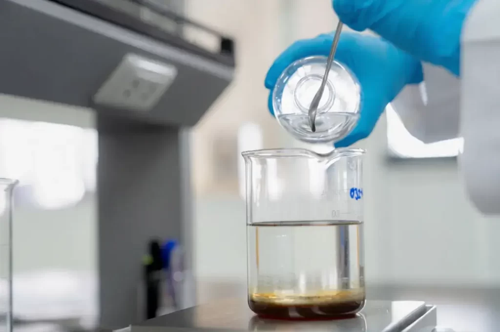 Close-up of a laboratory technician in blue gloves pouring liquid into a glass beaker sitting on a weighing pan.