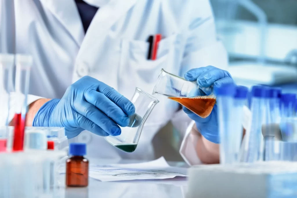 Close-up of a scientist in blue gloves pouring orange liquid from one beaker into another at a lab bench.