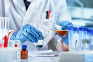 Close-up of a scientist in blue gloves pouring orange liquid from one beaker into another at a lab bench.