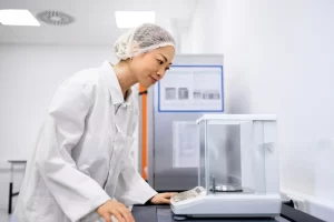 A female scientist in a lab coat and hairnet operates a precision lab balance in a clean laboratory.