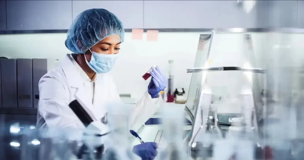 A female lab technician wearing a mask and hairnet examines a blood sample vial.