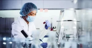 A female lab technician wearing a mask and hairnet examines a blood sample vial.