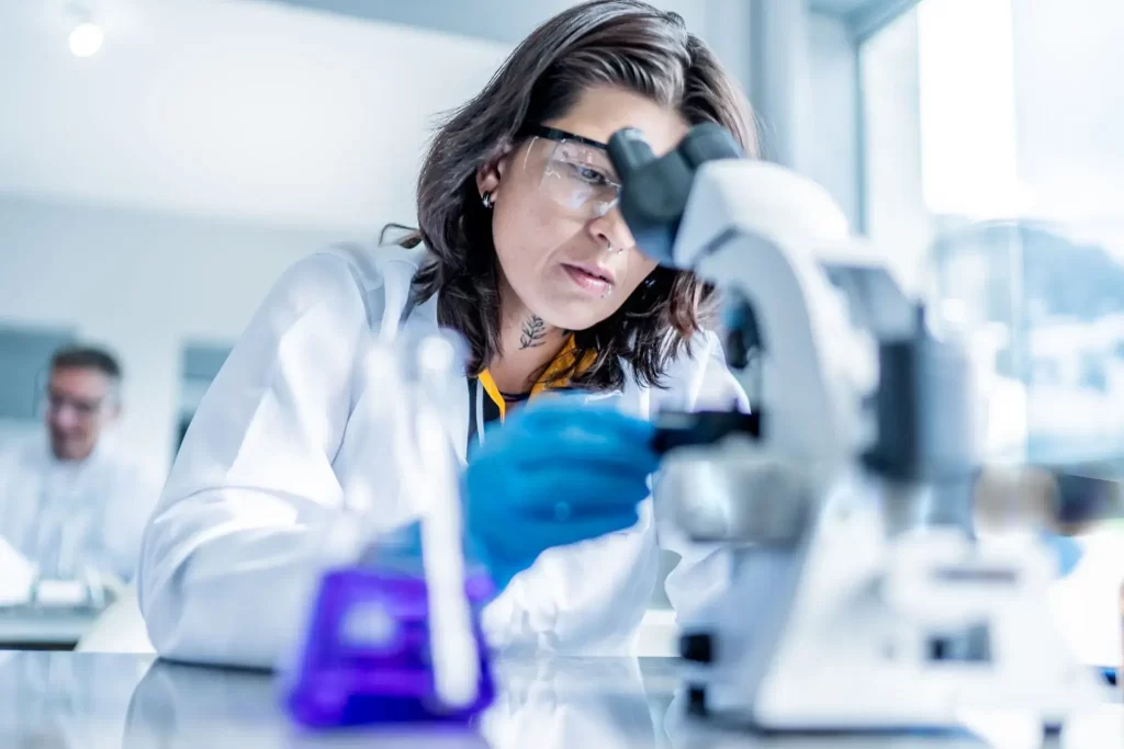 A female scientist in safety glasses and a lab coat carefully adjusting a modern optical microscope.