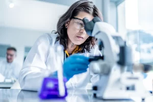 A female scientist in safety glasses and a lab coat carefully adjusting a modern optical microscope.