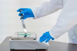 A technician in blue gloves uses a spatula to place chemical powder onto a top-loading balance.