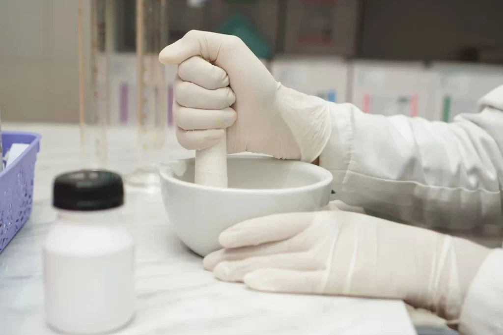 Pharmacist in gloves using a porcelain mortar and pestle to grind active ingredients for pharmaceutical compounding.