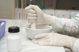 Pharmacist in gloves using a porcelain mortar and pestle to grind active ingredients for pharmaceutical compounding.