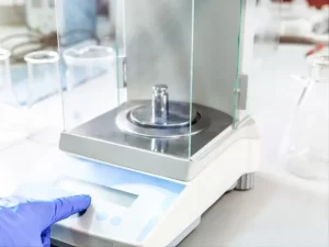 A laboratory technician in blue gloves operating the control panel of a precision analytical balance scale.