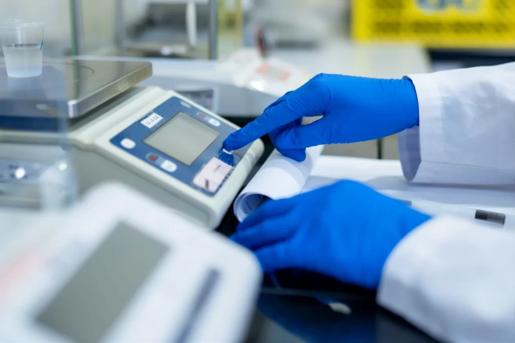 A laboratory technician in blue gloves taring a precision digital balance.