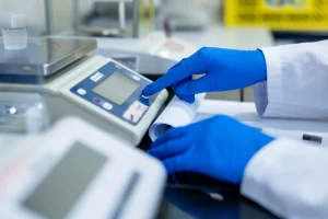 A laboratory technician in blue gloves taring a precision digital balance.
