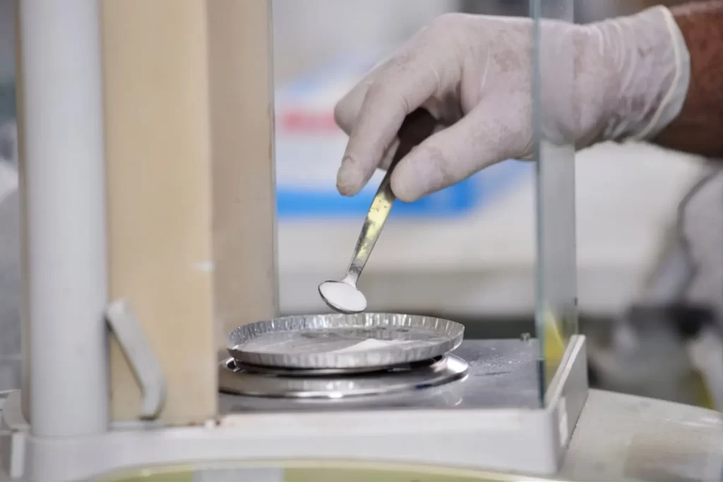Laboratory technician with gloved hand using a micro spatula to measure chemical powder inside an analytical balance.