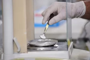 Laboratory technician with gloved hand using a micro spatula to measure chemical powder inside an analytical balance.