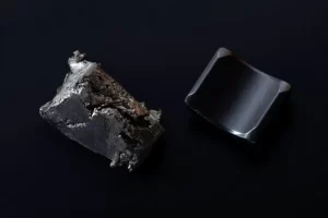 A raw magnetic mineral chunk next to a polished curved permanent magnet on a dark surface.