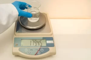 Laboratory technician placing a glass beaker with liquid on a precision balance pan.